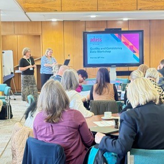 two people presenting at the front of a room. There are two tables with people sitting in chairs around them.